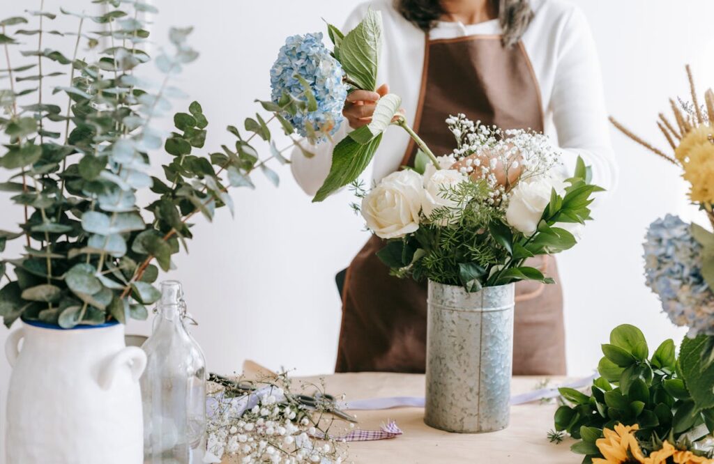 Crop unrecognizable female florist arranging hydrangea flowers into vase with roses while standing at table with assorted plants in room