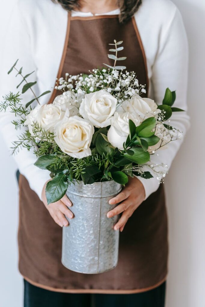 Crop unrecognizable female in brown colored apron standing with metal vase with bouquet of white roses in light room